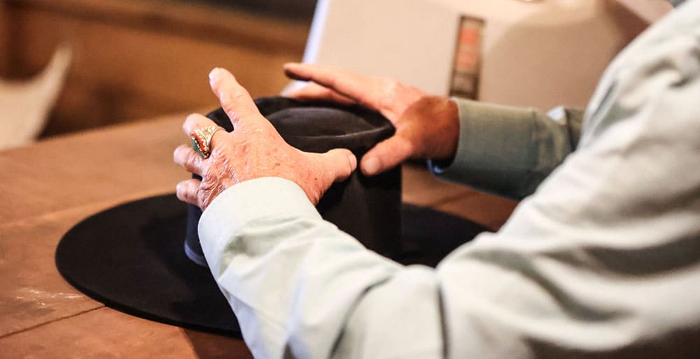 Old hands wearing a turquoise and red ring, shaping the crown of a black cowboy hat. 